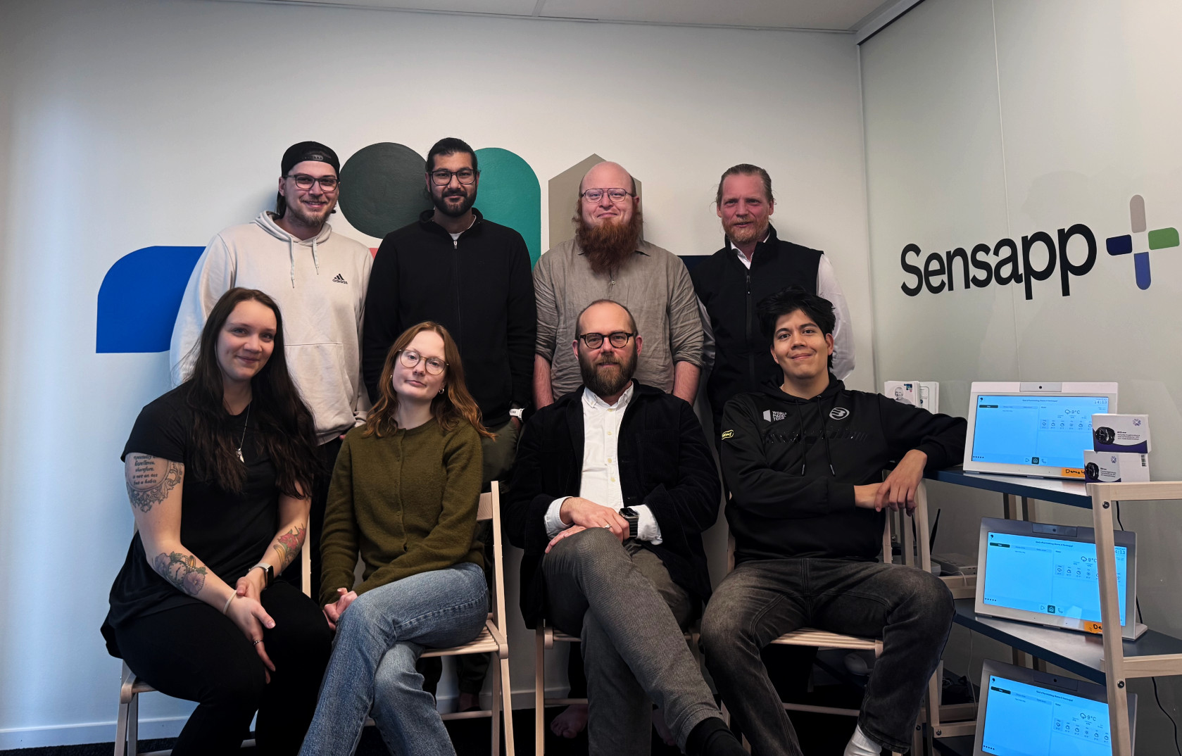 Sensapps team samlat framför företagslogotypen i de nya lokalerna i Göteborg – åtta medarbetare, fyra stående och fyra sittande, bredvid en hylla med Bubblan-enheter.